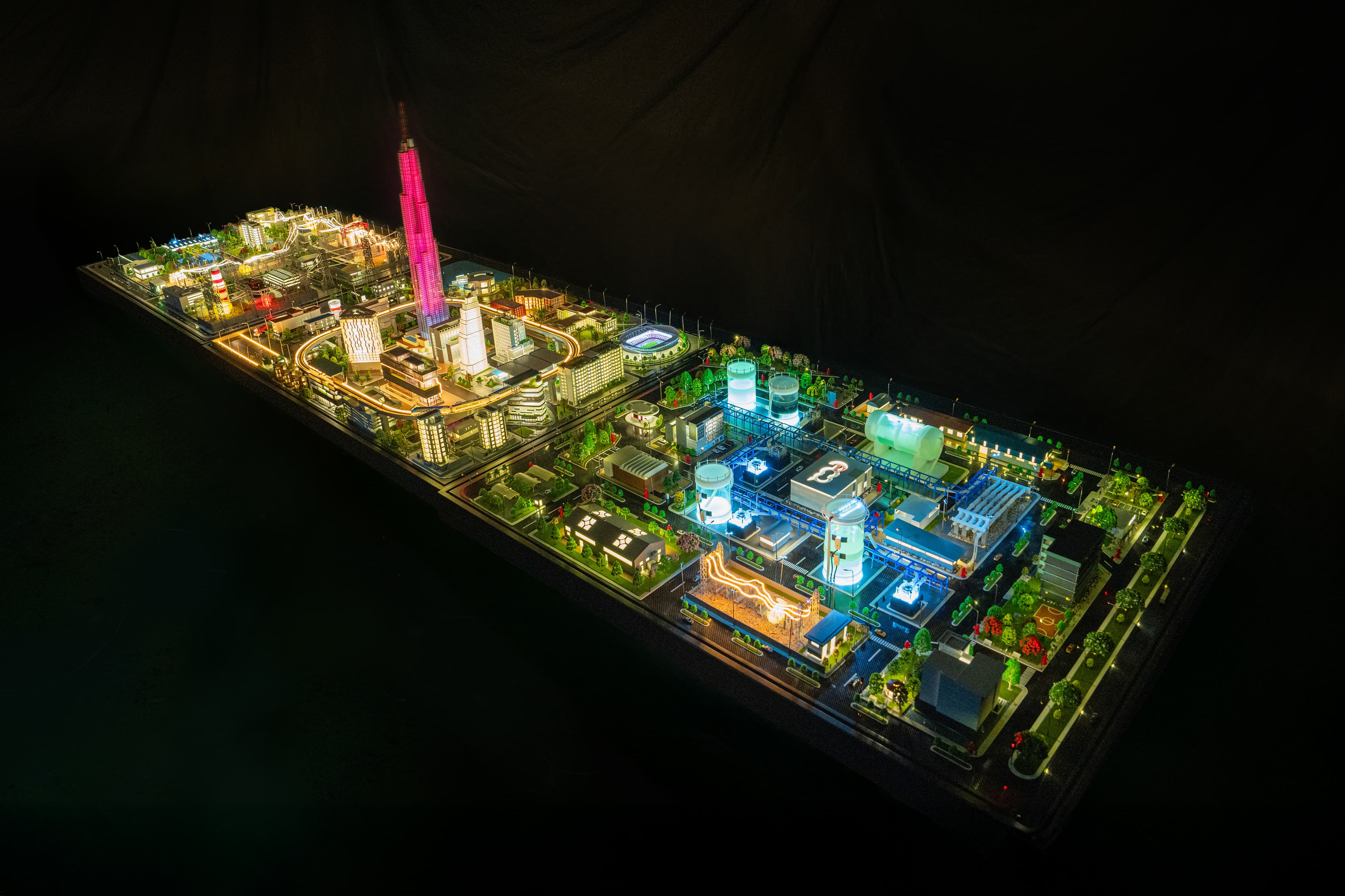 The range from above — the lit city district and its tower at one end, the process plant's tanks and blue pipe racks at the other, and the substation's lattice towers and live-lit lines along the front edge