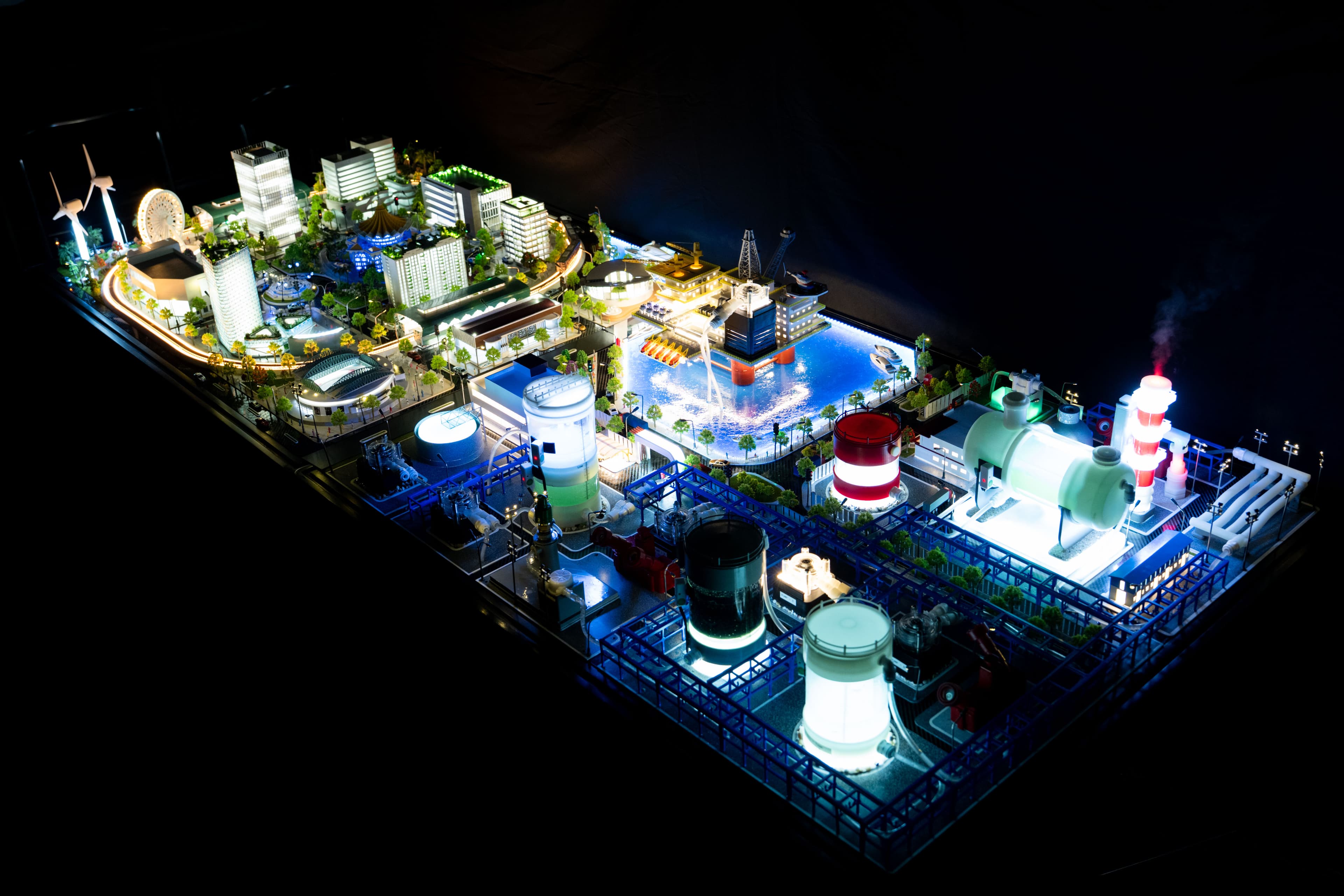 The physical OT Cyber Range model lit at night — a smart-city district, a harbour with an offshore platform, and the refinery skids running along the front of the table